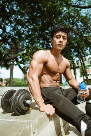 The shirtless muscular man sits holding a drinking bottle by the dumbbells after a hand muscle workout in the parkの写真素材