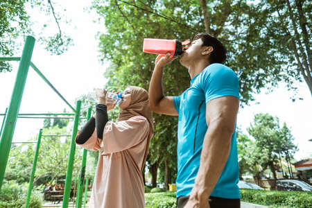 a muscular young man and a girl in a headscarf drinking with a bottle out of thirst during their outdoor sports break in the parkの写真素材