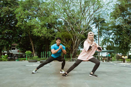 a young man and a veiled girl in gym clothes doing the leg warm-up movement together before exercising in the parkの写真素材