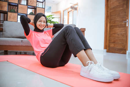 a girl in a hijab gym outfit smiles while doing a sit-up on a mat on the floor in the houseの写真素材