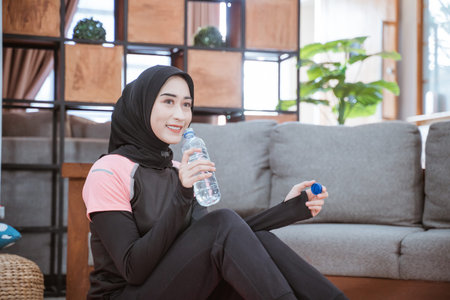 asian woman wearing a hijab gym outfit sits on the floor holding a drinking water bottle in the...
