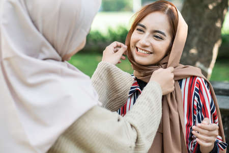 portrait of happy muslim asian woman fixing friend hijab while meeting outdoorの写真素材