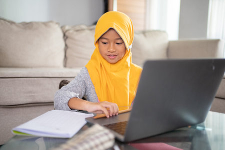happy muslim child wearing head scarf smiling while using laptop computer at home