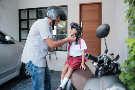 father taking his daughter to school by motorcycle in the morning. fasten the helmet. asian primary student wearing uniform going back to schoolの写真素材