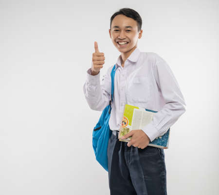 a smiling boy wears a school uniform and carries a backpack and book with thumbs up to the camera in an isolated backgroundの写真素材