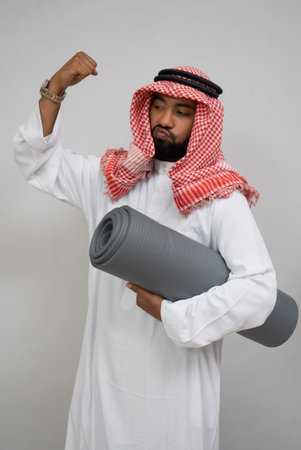 an Arab man wearing a turban feels the spirit of clenching his fist upward while carrying a mat on a white backgroundの写真素材
