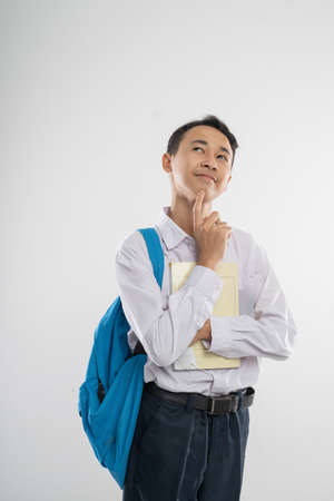 a boy in junior high school uniform smiling looking up when thinking gesture when holding a book and a backpack with copyspace on an isolated backgroundの写真素材