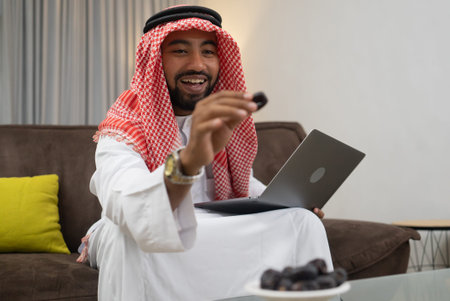 an arab businessman wearing a turban smiling while holding a date while working using a laptop in...