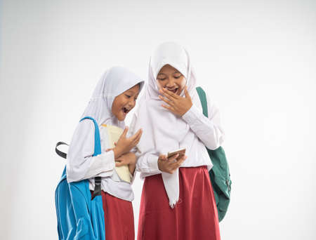 two veiled girls wearing elementary school uniforms using a mobile phone together with shocked expression when carrying backpack an isolated backgroundの写真素材