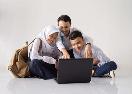 smiling three teenagers in junior high school uniforms sitting on the floor while using a laptop together against isolated backgroundの写真素材