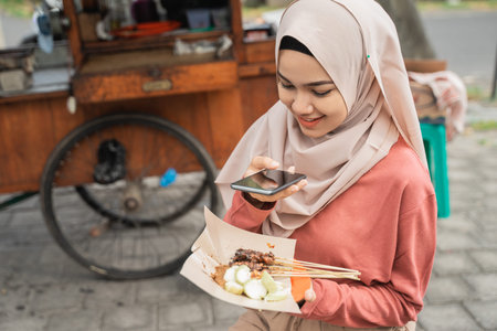 portrait of muslim sate ayam or chicken satay buyer taking photo of her food using smart phoneの写真素材