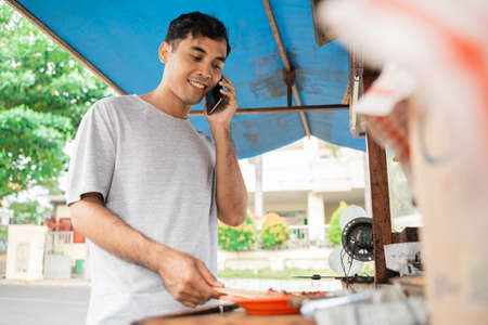 smiling indonesian sate ayam seller cooking the food while talking on a phoneの写真素材
