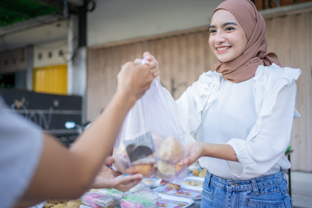 An asian girl in a veil gives takjil food orders in plastic bags to buyers at a roadside stallの写真素材