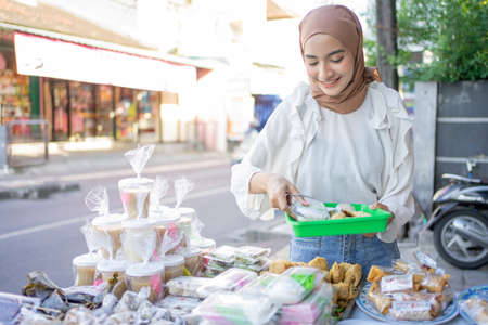 A beautiful girl in veil holding a plastic tray chooses various types of fritters to buy at a roadside stallの写真素材