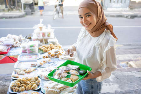 A beautiful girl wearing a headscarf uses tongs and carrying a plastic tray choosing snacks to buy at a roadside stallの写真素材