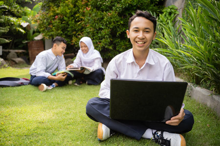 a teenager in a school uniform smiles while using a laptop sitting on the grass in the garden with...