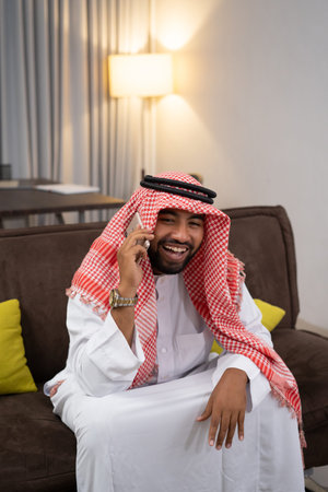 Arab businessman in a turban laughing while using a cellphone while sitting on the sofa with a floor lamp and curtains as a backgroundの写真素材