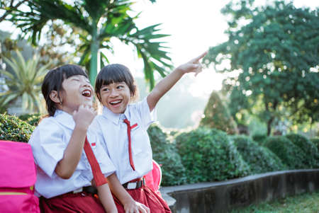 cute two asian student in uniform pointing away while sitting togehter in the parkの写真素材