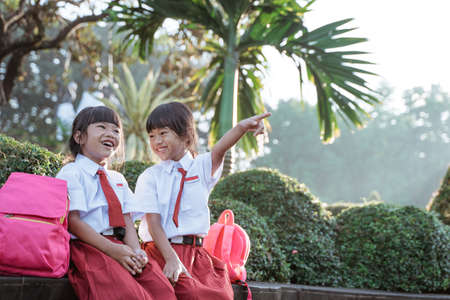 cute two asian student in uniform pointing away while sitting togehter in the parkの写真素材
