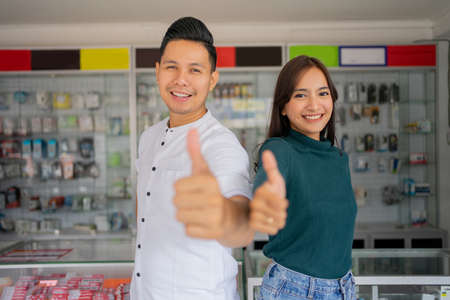 close up of a handsome young man and beautiful young woman with thumbs up standing on a smartphone shop interior design backgroundの写真素材