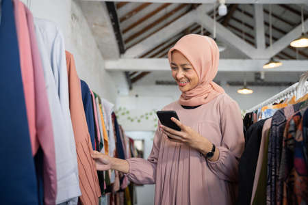 a beautiful girl in a hijab choosing clothes while holding a smartphone in a boutique shopの写真素材