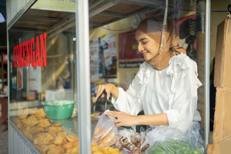 a beautiful girl wearing a hijab while using tongs chooses fried food the buyer orders on a roadside cart. Gorengan is indonesian word mean fried snackの写真素材