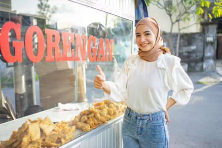 a beautiful girl in a veil with thumbs up selling various kinds of fried food using a cart on the side of the road. Gorengan is indonesian word mean fried snackの写真素材