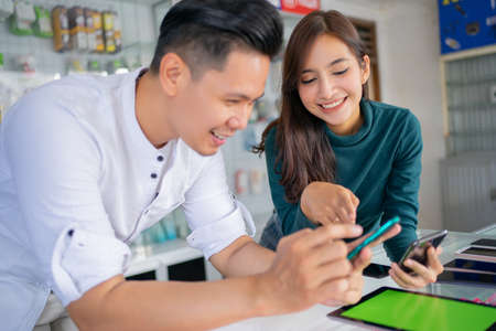 a beautiful girl smiles while pointing at the mobile phone screen of a male shop assistant on the background of a smartphone shopの写真素材