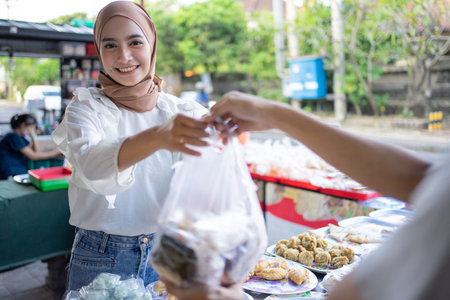 A beautiful girl wearing a headscarf selling a food stall on the side of the road gives takjil food to a buyer at the stallの写真素材