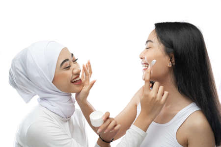 Smiling asian muslim bestfriend applying face cream with finger hands in front of camera with gray backgroundの写真素材