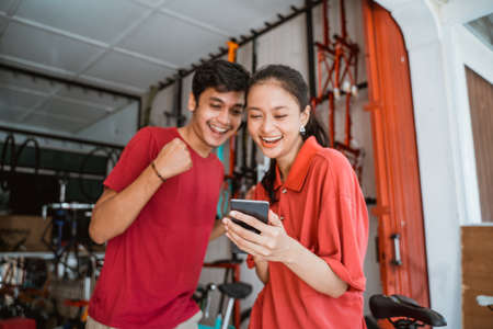excited asian young couples at bike shop suprised while looking at phoneの写真素材