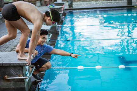teenage swimmer standing on block board and preparing to jump and swim with a coach giving instructionsの写真素材