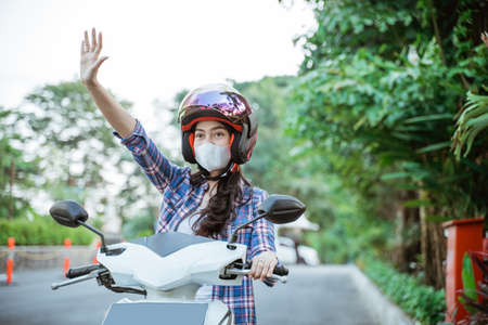 Asian woman wearing helmet and mask waving while riding motorbikeの写真素材