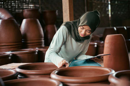 asian veiled woman looking closely at earthenware while holding tabletの写真素材