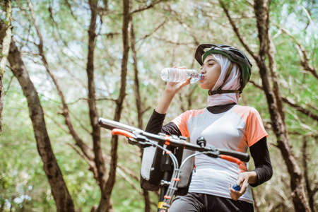 Woman in hijab drinking mineral water bottle when thirsty while cycling on outdoor backgroundの写真素材