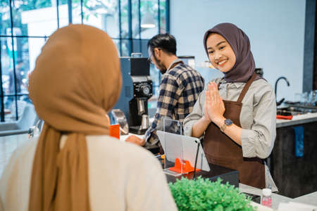 beautiful asian muslim cafe worker welcoming customer at her shopの写真素材