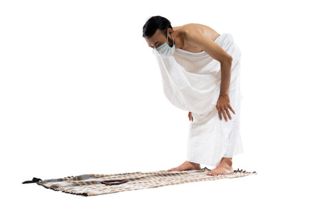 Man in mask wearing ihram clothes praying with prayer rug while bowing on isolated background