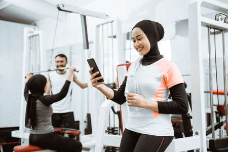 beautiful muslim sporty woman taking a break after exercising at the gym sitting on the floor and using phoneの写真素材
