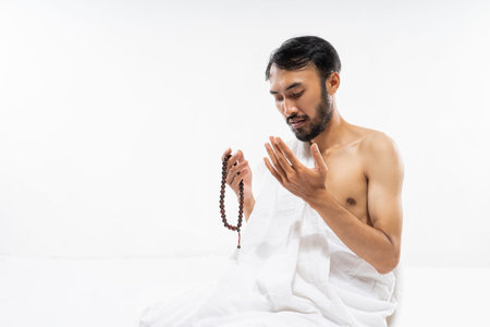 Asian bearded man wearing ihram clothes praying with prayer rug on isolated background