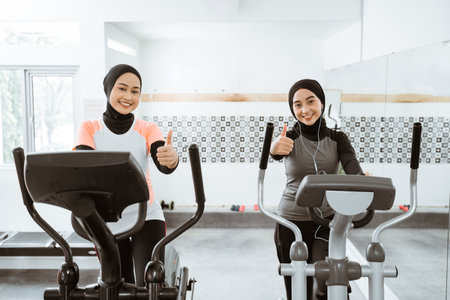 two beautiful woman with hijab at the gym using static elliptical cycle machine and showing thumb upの写真素材