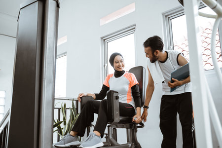 Personal trainer helping woman with hijab working with leg weight machine at the gym