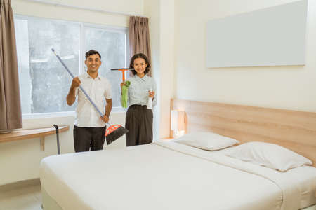 Two cleaning staff holding cleaning tools standing near bed in hotel roomの写真素材