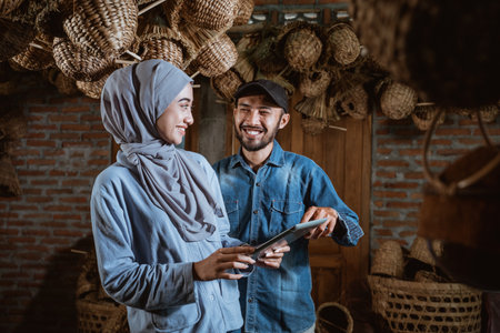 male and female entrepreneurs in hijab chatting using a tablet against a water hyacinth handicraft production house in the backgroundの写真素材