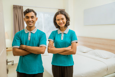 two janitors wearing turquoise uniforms smiling while standing with their arms crossed in a hotel roomの写真素材