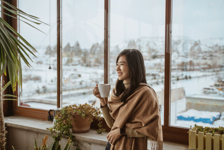 beautiful asian woman enjoy coffee morning standing next to a big window with beautiful view of snowy landscape in cappadociaの写真素材