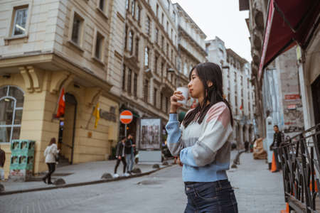 woman wearing sweater drinking her coffee while standing in city centre surrounded by classical buildingの写真素材