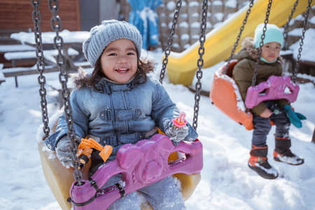 close up portrait of a cute little girl smiling while swinging in the playground full snow in winterの写真素材