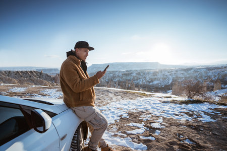 man using his mobile phone at the top of the hill while leaning against his car with beautiful view...