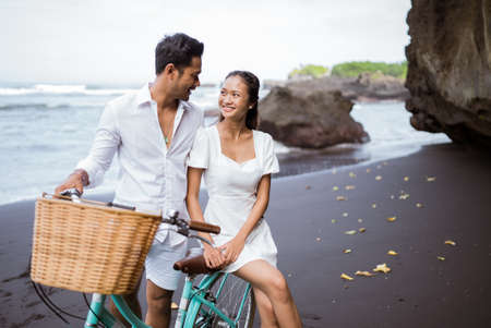 happy asian young couple riding bicycles on black sandy beachの写真素材