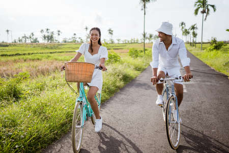 couple leisure and lifestyle concept - happy young asian man and woman riding bicycles along road in summerの写真素材